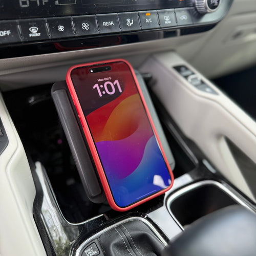 A red smartphone in a red case rests on a black wireless charging pad placed in a car's center console. The console has beige leather trim and a black climate control panel.