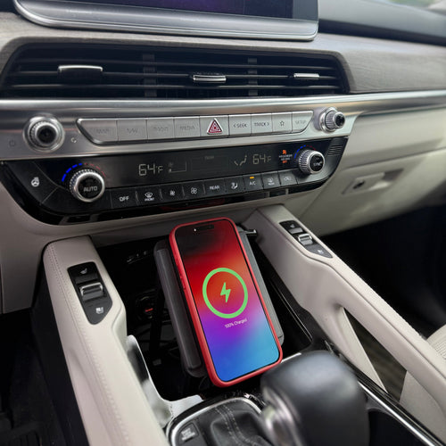 A smartphone with a red protective case lies on a wireless charging pad installed in a car's center console. The car interior features beige leather trim and a black dashboard infotainment screen.