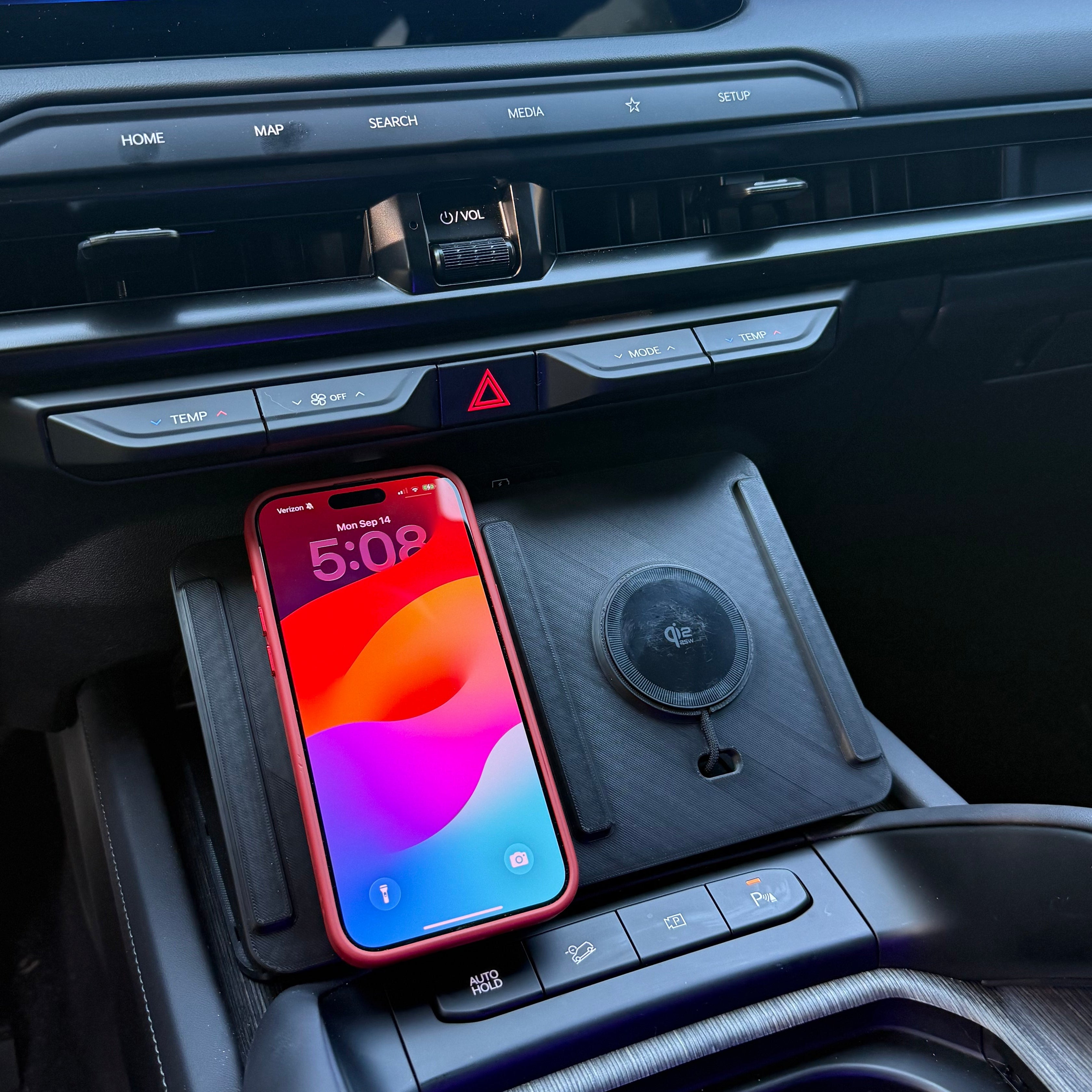 A smartphone in a red case rests on a black wireless charging pad inside a car's center console, with the car's infotainment screen and dashboard visible in the background.