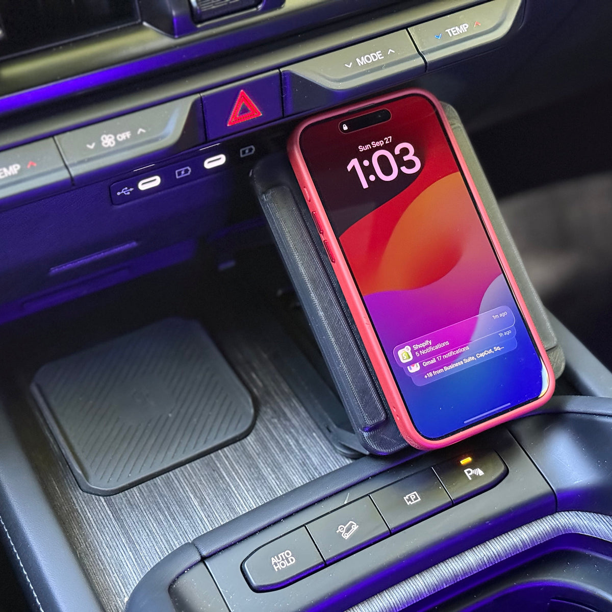 A red smartphone in a red protective case rests on a black wireless charging pad inside a car's center console.