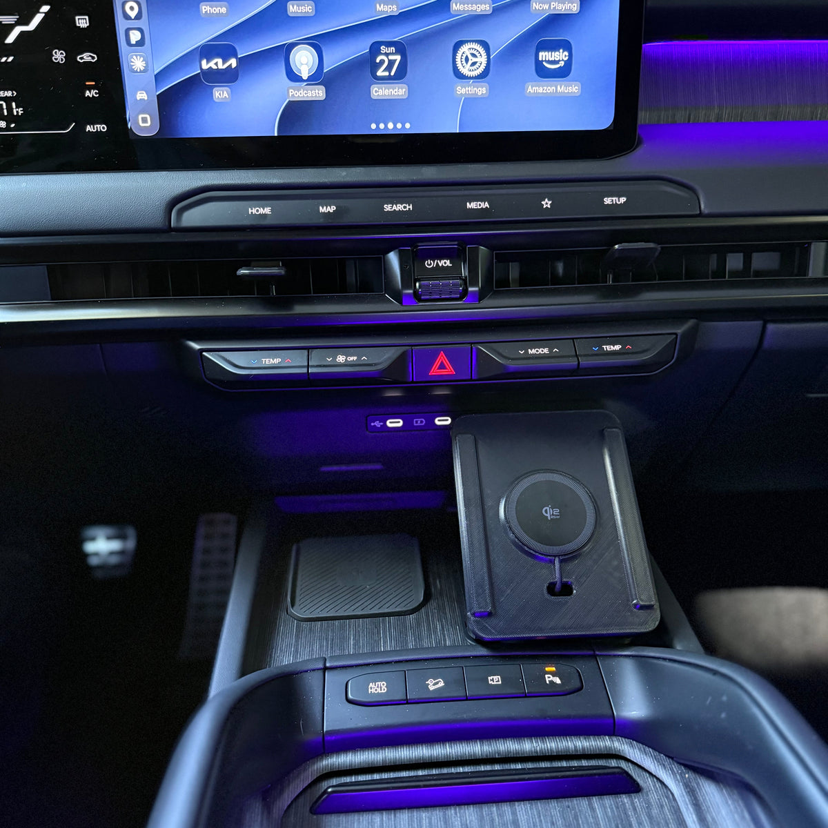 Close-up of a car interior console with a large illuminated touchscreen, center console controls, a wireless charging pad, and ambient purple lighting.
